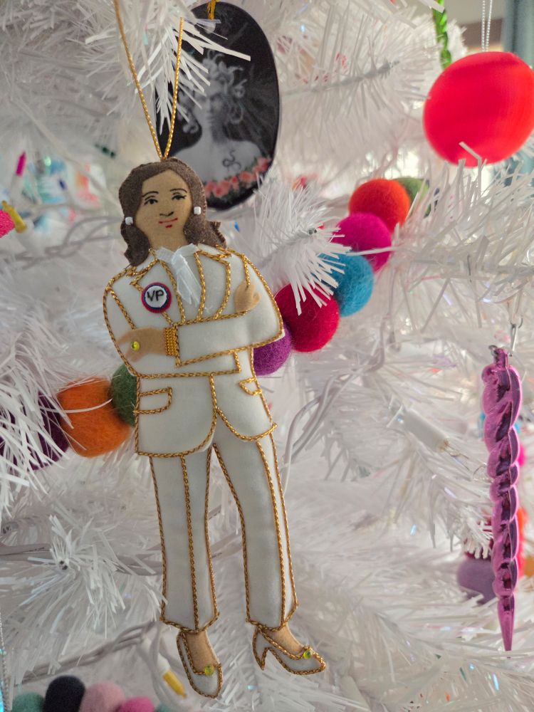 White faux Christmas tree with felt ball garland setting off a handmade Kamala Harris ornament where she's wearing a white pant suit and pearl earrings. A Medusa oval-shaped ceramic ornament admires her from the background. 