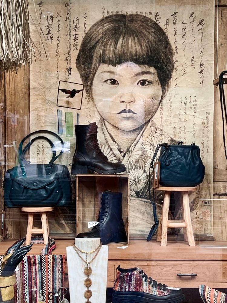 Blick in ein Schaufenster in dem Schuhe und Handtaschen, die auf kleinen Hockern platziert stehen, ausgestellt sind. Der Hintergrund des Fensters besteht aus einer großen Zeichnung eines asiatisch aussehenden Kindes und asiatischen Schriftzeichen. 