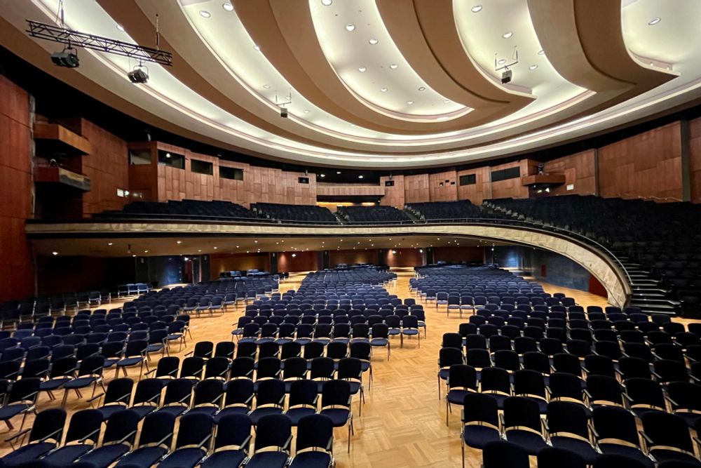 Auf einem hellen Parkett stehen viele reihenweise blaue Polsterstühle. Im hinteren Bereich führt eine geschwungene Treppe zu einer großen Empore. Die geschwungene Rückwand des Saales hat eine rotbraune Holzverkleidung. Die Decke ist mit geschwungenen Lichtelementen bedeckt, die gegenläufig zu den Kurven der Empore verlaufen. 