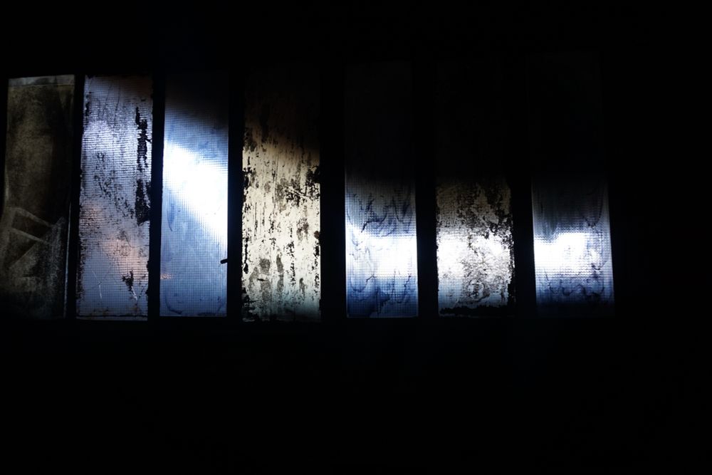 Blick auf eine schwarze Wand, in der eine Reihe Fenster aus Drahtglas sind. Die Scheiben sind teilweise zerbrochen und leuchten durch das dahinter scheinende Licht in Blau-und Grautönen. 