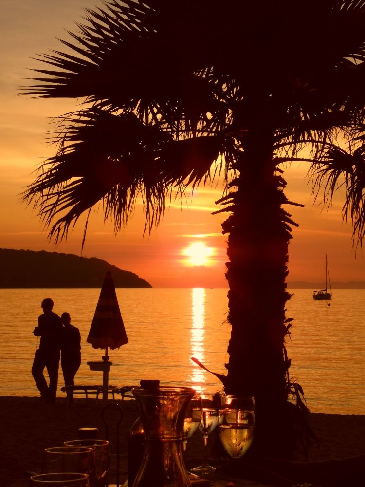 Blick auf einen Sonnenuntergang am Meer mit orangefarbenem Himmel und Meer. Im Vordergrund sind am Sandstrand im Gegenlicht die dunklen Silhouetten einer Palme und eines Sonnenschirms zu sehen, daneben die eines Paares, das nebeneinander geht. Draußen auf dem Meer sind die Umrisse eines Segelbootes zu erkennen. Ganz im Vordergrund stehen eine Karaffe und Weingläser, durch die das Sonnenlicht scheint und in denen sich die Palme spiegelt. 