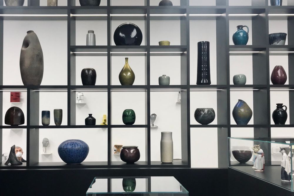 Blick auf ein großes, dunkelgraues Regal vor einer weißen Wand. Die Fächer sind mit wenigen Ausnahmen quadratisch angelegt. In jedem Fach steht eine Vase. Es gibt unterschiedliche Formen und Größen, zum Beispiel bauchig, schmal oder asymmetrisch. Die Farben sind überwiegend dunkel, von schwarz über violett , braun, rot, bis blau oder grün. Die Regalfächer sind beleuchtet und die Glasuren der Vasen glänzen.