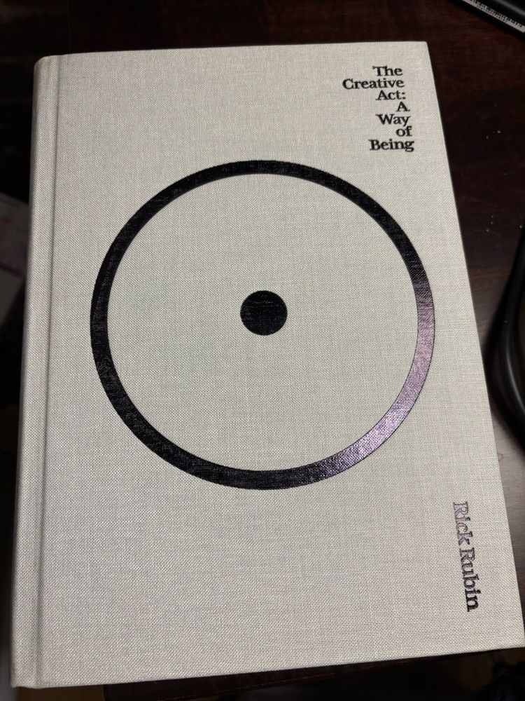 Hardcover copy of The Creative Act: A Way of Being by Rick Rubin. The book is a pale, desaturated gray/green. On the cover, there is a massive black embossed circle with a dot in the center. The title and author are also embossed black and tucked away to the right. 