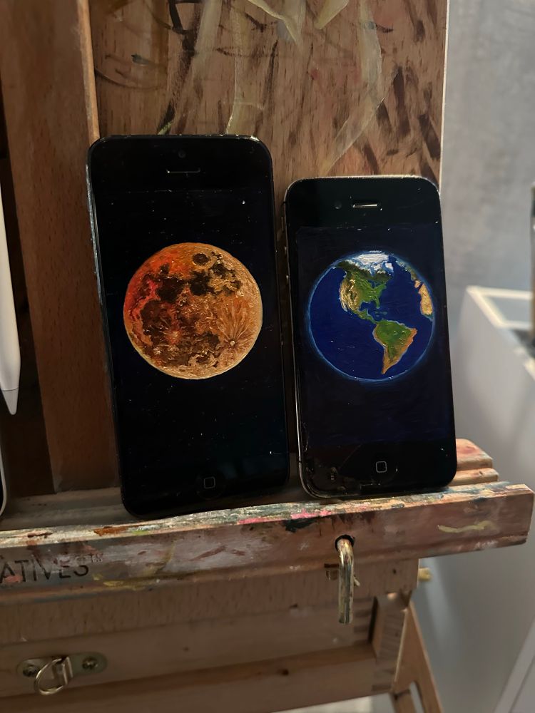 two iPhone leaning against an easel - both have small oil paintings on their surfaces. the left shows a strawberry moon and the right shows earth facing north and South America. 
