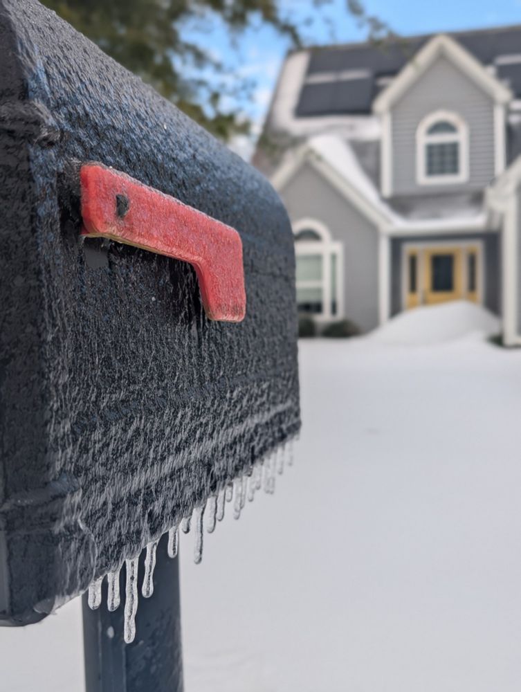 Frozen mailbox covered in ice with small icicles (in focus) with a snow covered yard and front of a house in the background (soft focus).