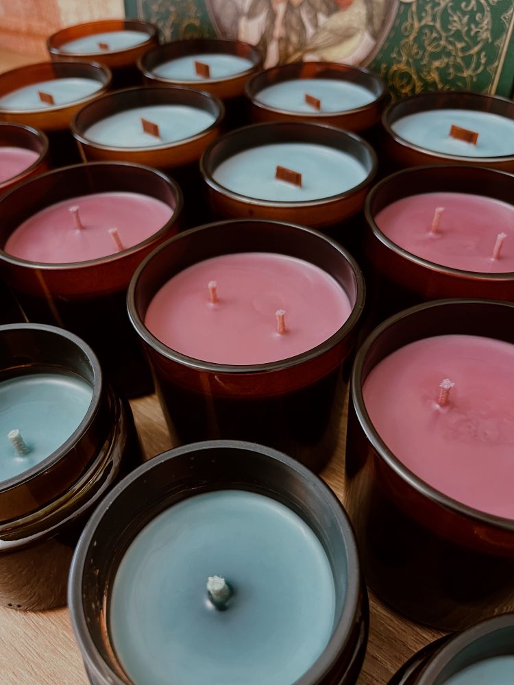 A foto possui várias velas, de diversas cores, todos os frascos de vidro âmbar. Tem velas de pavio de madeira, pavio duplo de algodão e pavio único de algodão. 