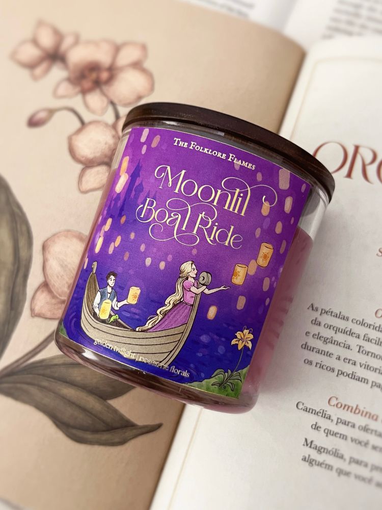 A foto possui uma vela deitada em cima de um livro aberto, com desenho de flores. A vela possui um frasco transparente e uma cor vinho, com tampa de madeira. Seu nome é “Moonlit Boat Ride”, com uma ilustração inspirada no filme da Disney Enrolados.