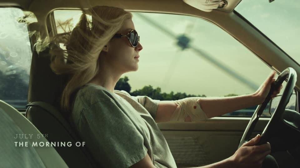rosamund pike driving in her car with the windows open and sunglasses on, a bright day, you can see the bandage on her arm seeping blood a little, and a title on screen "july 5th: the morning of"