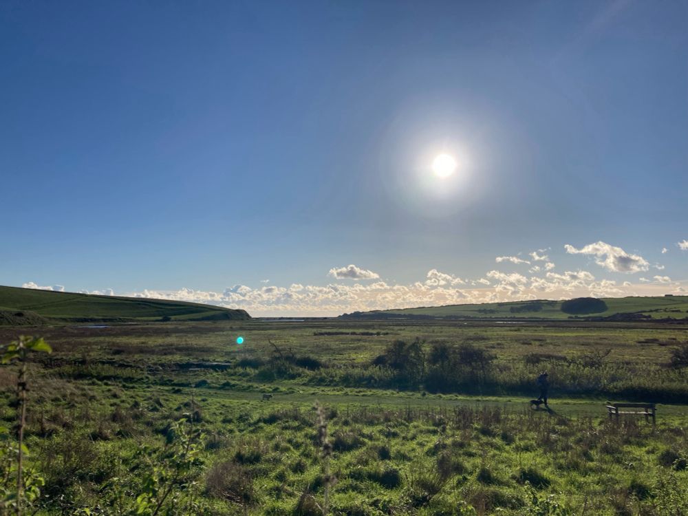 Photograph of valley, with ox bow river just visible, blue sky, sun and light clouds on horizon.