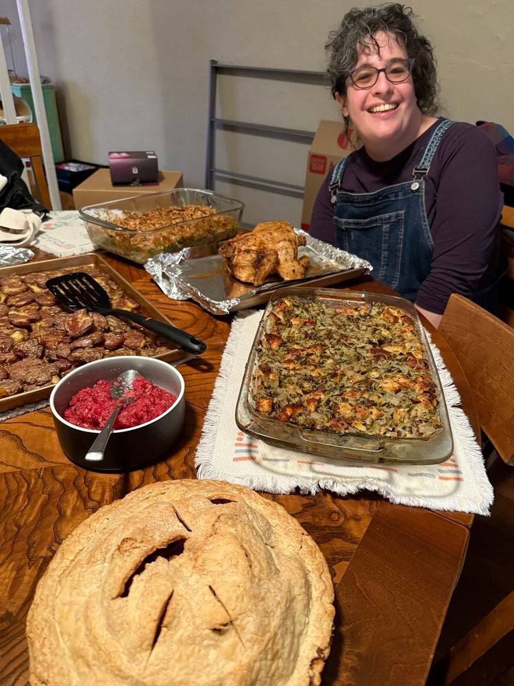 a salt & pepper haired woman in blue overalls & a purple shirt beams behind a spread of thanksgiving treats including a store bought rotisserie chicken, a pan of stuffing, a pan of green bean casserole, a baking sheet of smashed potatoes, a bowl of cranberry relish and a homemade apple pie