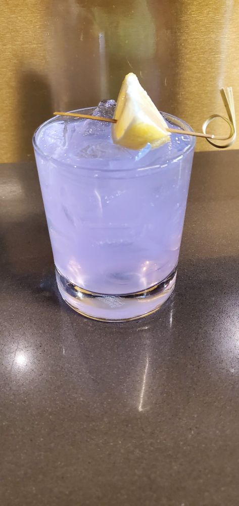A lavender colored cocktail with a lemon, called Iris You Well