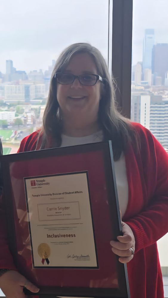 Carrie, a woman with medium length brown hair and glasses, wearing a red sweater and white shirt, stands at a window in front of the Philadelphia skyline.  She is smiling and holding a large framed certificate with a red border. Her name and the word "Inclusiveness" can be read; the other words are too small. 