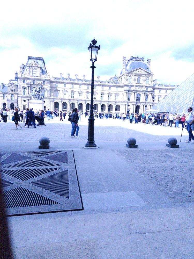 Courtyard outside of the louvre. The glass pyramid is to the right. There are many people waiting in lines. The sun is bright.