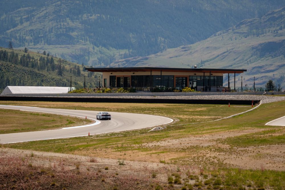 A white ford mustang taking the left hand turn 16 at area 27. The clubhouse and tree lined hills behind. 