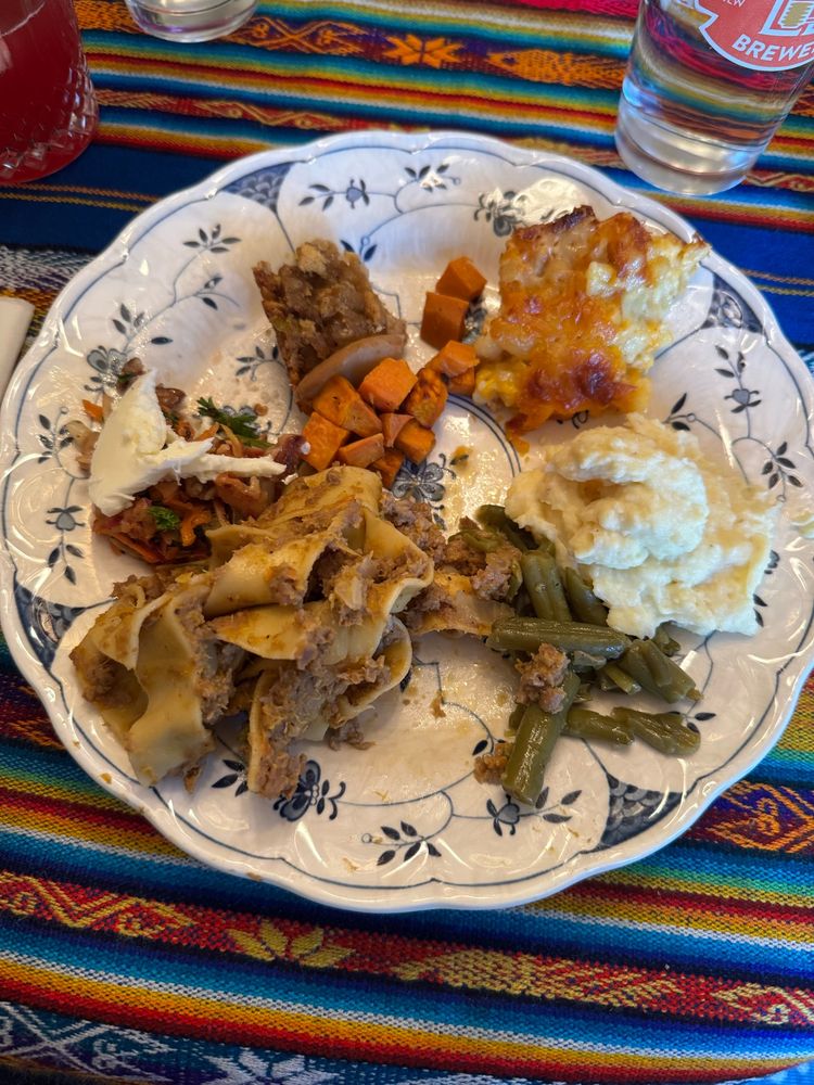 My plate on Thanksgiving, partially eaten, with pumpkin bolognese, carrot salad with burrata, stuffing, sweet potato, Mac and cheese, green beans, and mashed potatoes on top of a multicolor table runner 