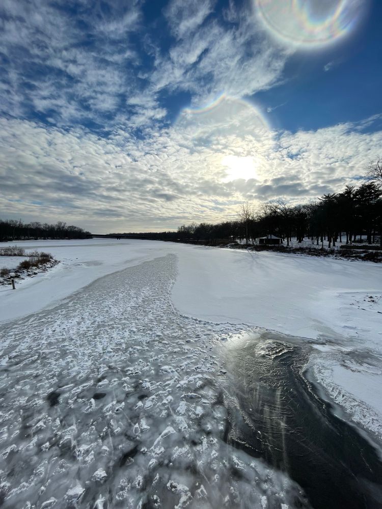 Frozen river flowing towards the afternoon sun. Sky is blue with a flock of clouds gathered heavily at the horizon line. Darkened trees line the riverbanks. 