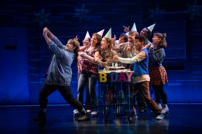 A crowd of people wearing ice skates and cone-shaped party hats pose for a group selfie. [The cast of Kimberly Akimbo. Photo by Joan Marcus]