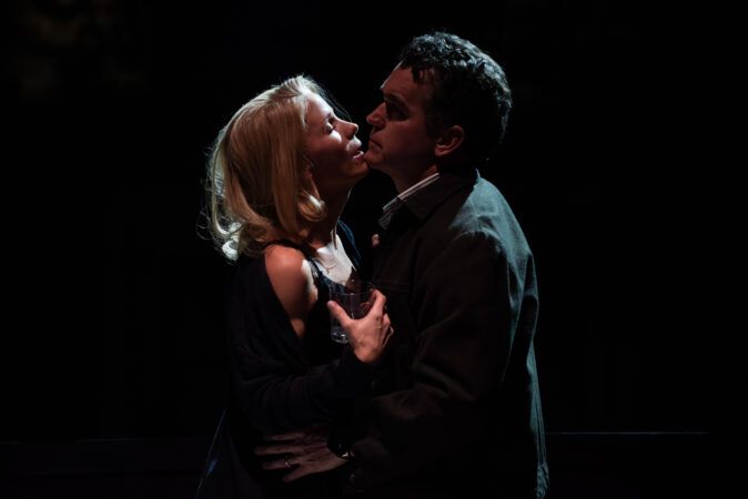 Against a black background, in harsh side-lighting, a white man and woman are almost embracing, but for the tumbler of gin in her hand, and his face turning away from a kiss. [Photo: Kelli O'Hara and Brian d'Arcy James as Kirsten and Joe in Days of Wine and Roses. Photo by Ahron R. Foster]