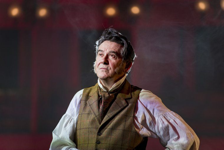 Adrian Lukis as George Wickham. Photo by James Findlay. Image: a white man in his sixties, with muttonchop sideburns and in early Victorian Era wardrobe, looks up and to the left, as cigar smoke wafts around behind him.