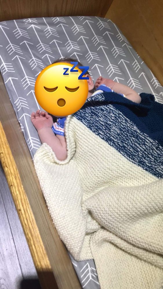 A bay blue and white blanket on top of a sleeping baby. The blanket has a marked section in the middle. The baby is lying with their arms out on a wooden crib. 