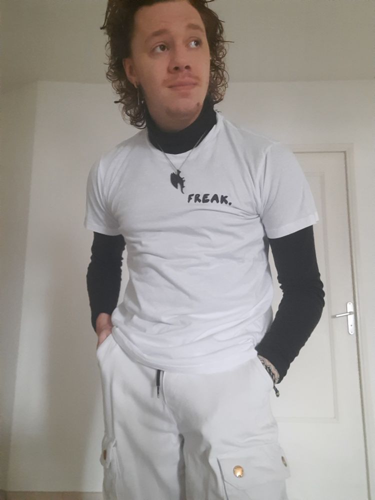 Beautiful person with white cargo pants, a white t-shirt with "freak." written on the front over a black turtleneck
The background is a white wall