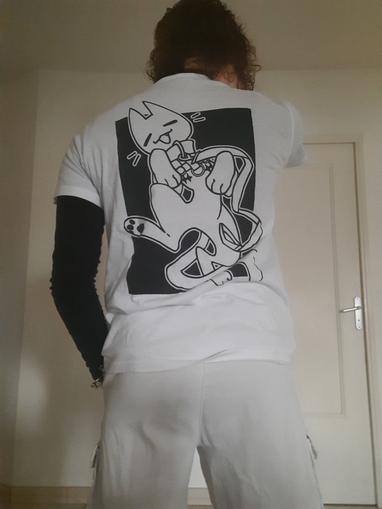 Same person but turning their back to the camera, showing the back art of the t-shirt a cartoon cat with a leash and top surgery scars