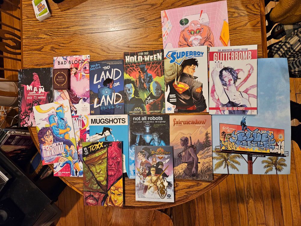 An overhead shot of comics and art. 
Maw
Fourth Dimensional parts 1 & 2
Bad Blood deck + comic
This Land is Our Land
Mugshots
Toxx vol. 1
Not All Robots
King Arthur & The Knights of Justice
Star Trek Holo-Ween
Superboy & The Man of Tomorrow
Fairmeadow
Glitterbomb
Print of Master Roshi with magazines
Print of Blue Beetle on billboard