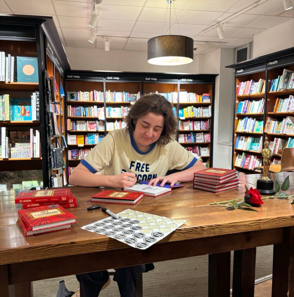 Emily is sat at a Waterstones table signing copies of her book Icons of Cinema: Baz Luhrmann. She’s wearing a white T-shirt with a blue graphic reading ‘free popcorn’ 