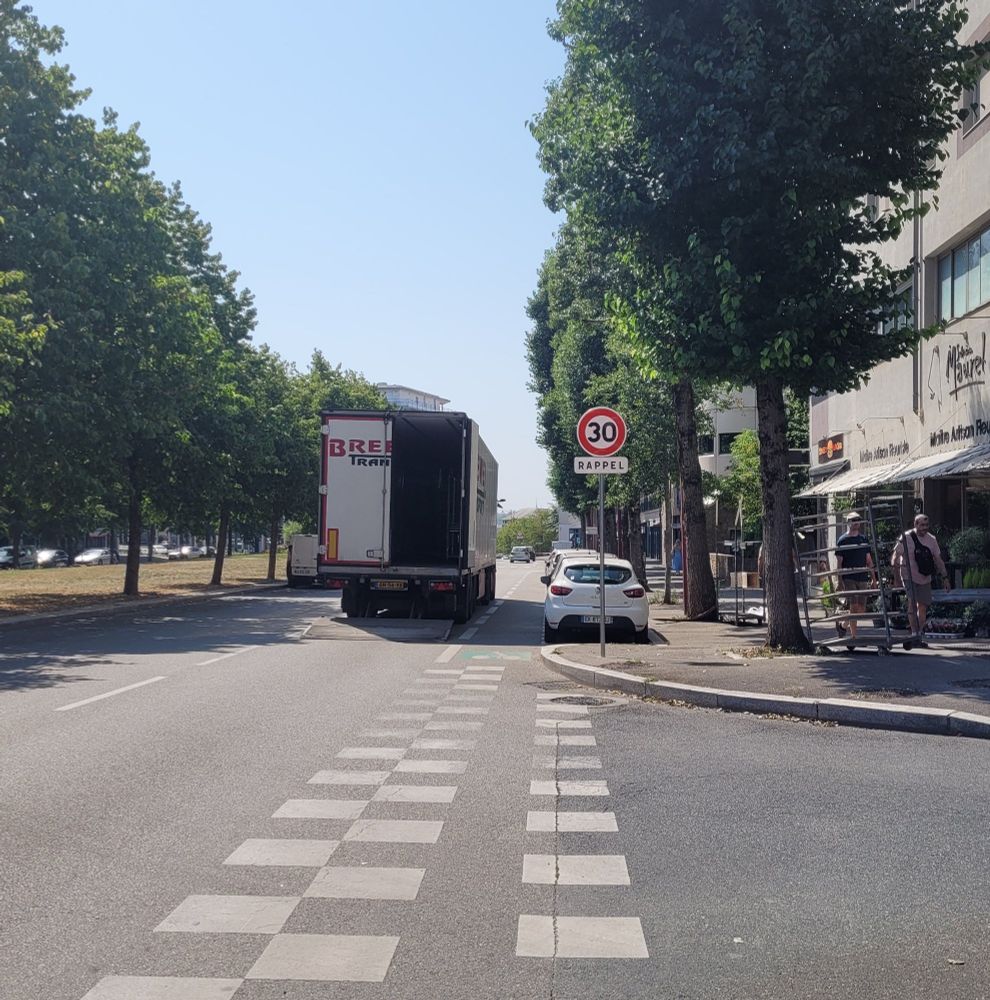 De gauche à droite : une voie de stationnement vide, une voie de circulation, une deuxième voie de circulation où stationne un camion, une bande cyclable, une voie de stationnement occupée, un large trottoir.