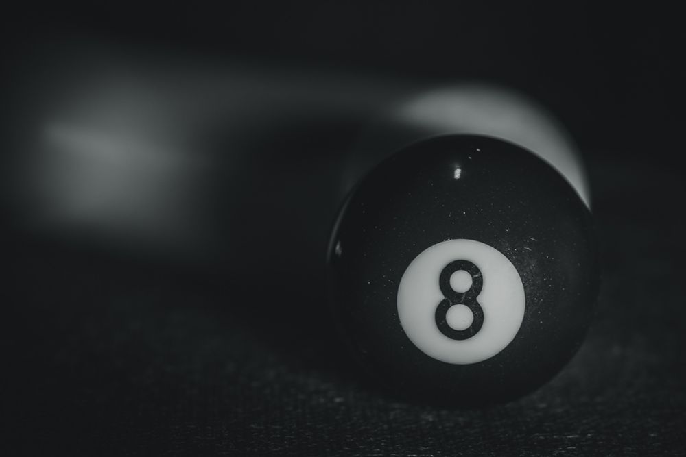 Eine hochglänzende, schwarze Billardkugel mit der weißen Zahl Acht in einer kontrastreichen Schwarz-Weiß-Aufnahme. Die Kugel ist scharf fokussiert und liegt auf dunklem Filz. Hinter ihr deutet ein länglicher, verschwommener Schweif die rasante Bewegung der weißen Kuge anl – die Acht im Moment der Entscheidung. Eine sehr dynamische, hoffentlich  meditative Szene.