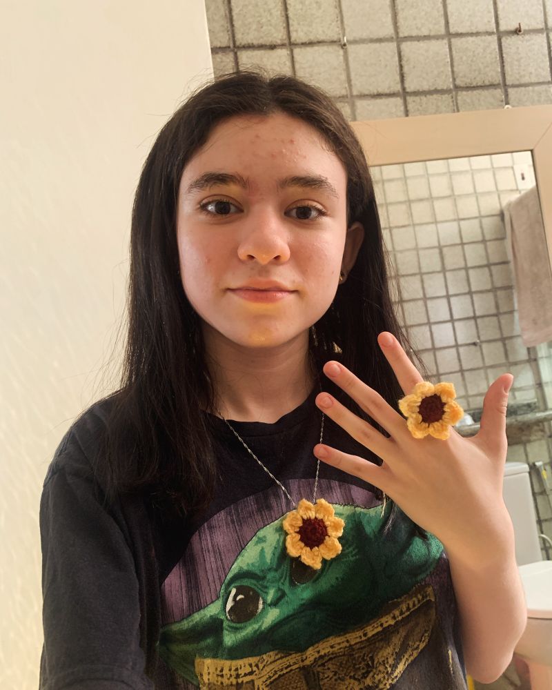 Luíza, uma menina branca de cabelos castanho escuro e olhos da mesma cor, está usando um colar de girassol de crochê e está mostrando a própria mão de modo que é possível ver seu anel grande e alegre de girassol de crochê.