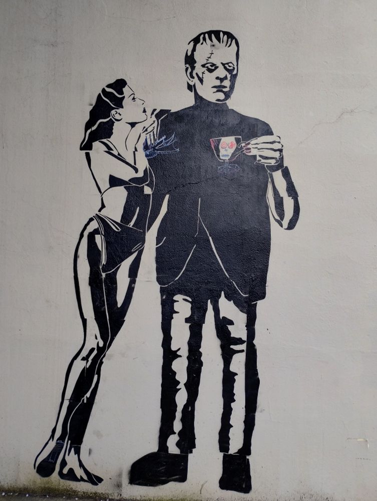 Spray painted stencil of a woman in bikini leaning on Frankenstein's shoulder while he holds a tea cup with a skull on it. 