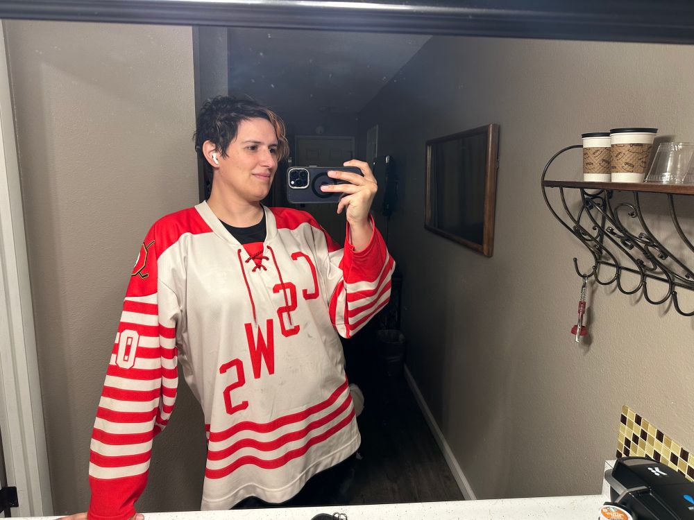 Ember stands in front of a mirror in a hotel room, taking a selfie in the mirror. Shi is wearing an oversized goalie hockey jersey in white with red stripes that make it look like a candy cane. The letters CSWS are diagonally across the chest, reversed cause of the mirror, and there’s red drawstrings hanging from the neckline.