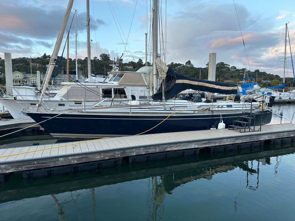 A sailboat with dark blue sides and gold trim tied up in a marina, all her sails and lines stowed. The sail cover for the mainsail is a matching dark blue with a gold stripe.