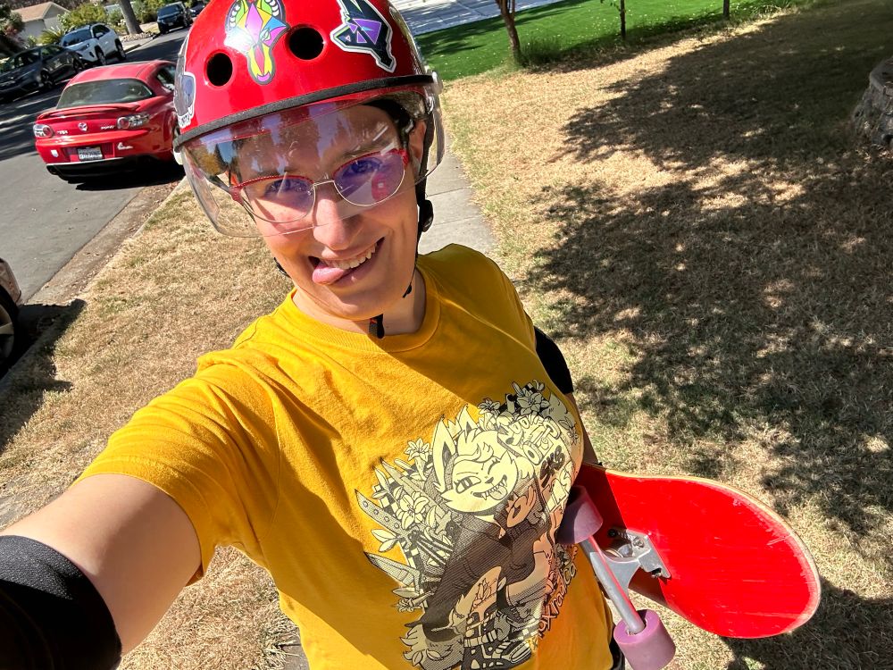 Selfie in a front yard. Ember is wearing a yellow shirt that has a wolf girl skateboarder on it, whose board says “skate fast eat ass” on the bottom. Ember is wearing a red helmet with a visor and stickers and carrying a red skateboard with purple wheels.