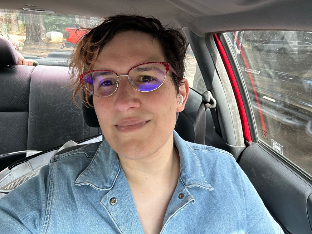 Selfie in a car. Ember is wearing a denim jumpsuit with the collar open, and looks exhausted. Hir hair is messy and sweaty, but shi has a genuine smile.