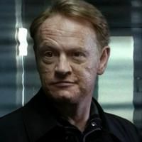A close up of a middle aged white man with reddish hair wearing a black jacket and staring slightly to the left with a serious look. David Robert Jones from Fringe.