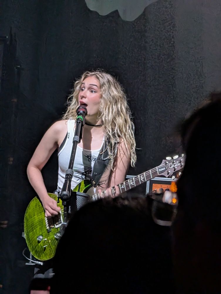 A young blonde woman wearing a white tank top playing a lime green guitar and singing at a microphone as she looks off to the left.