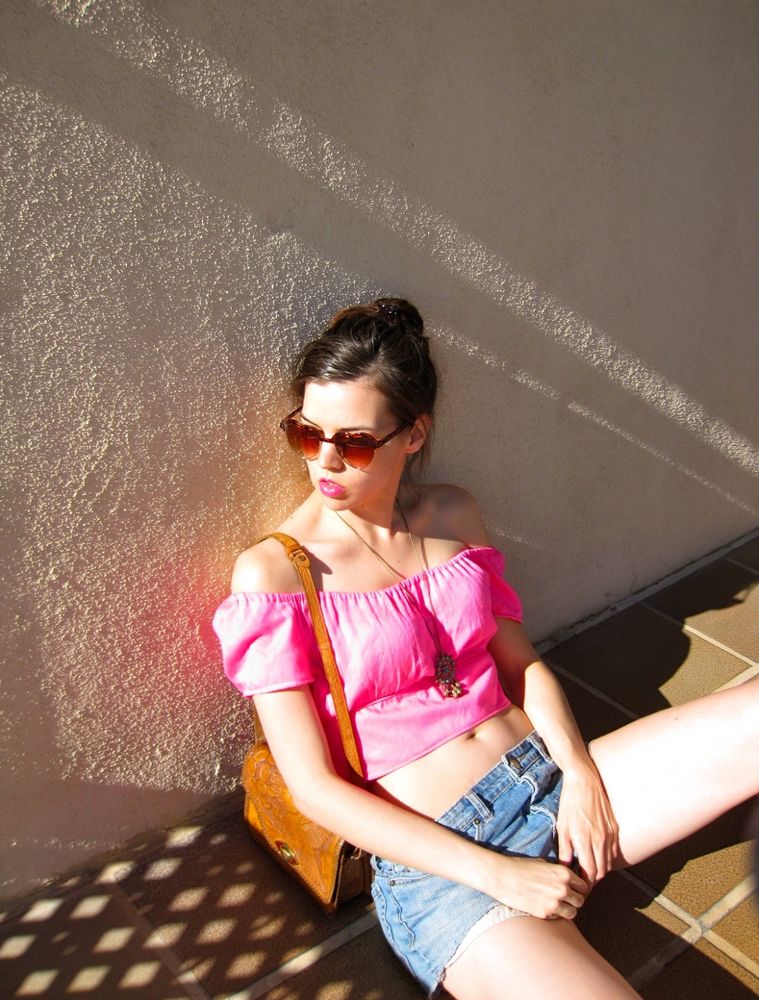 White brunette femme sitting against a sunlit wall in a pink crop top and jean shorts