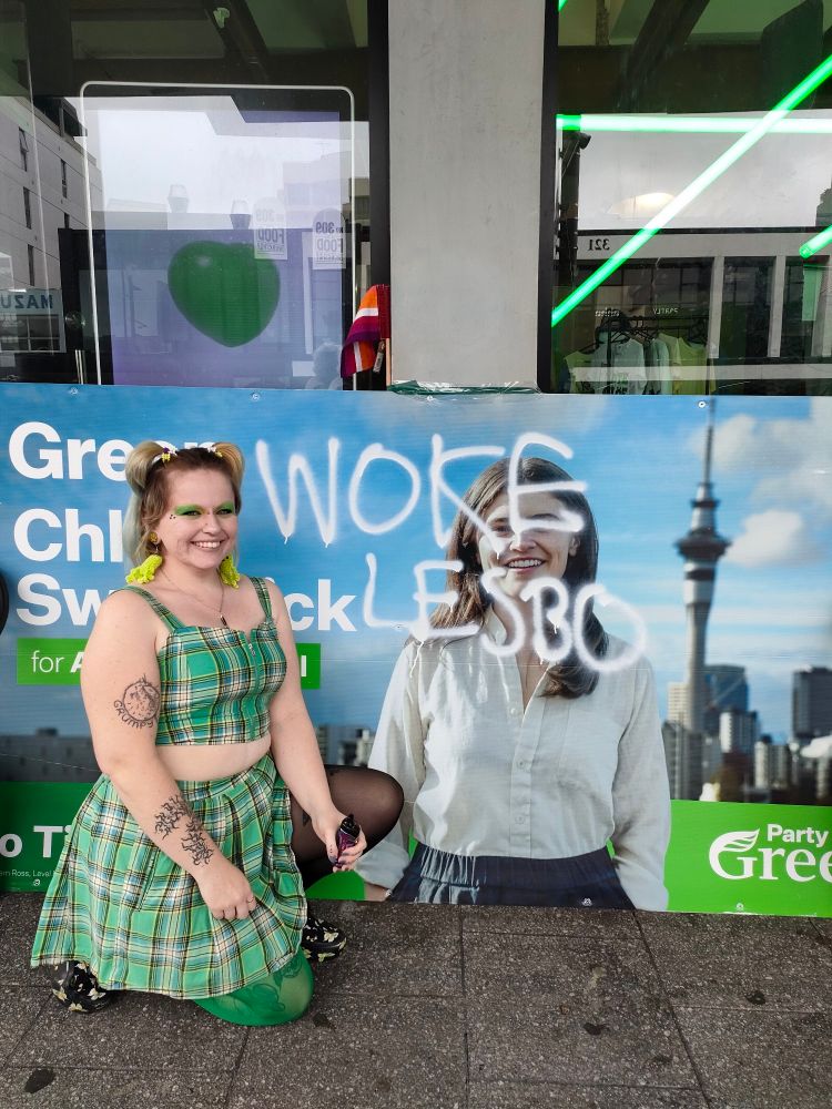 Tempest in front of the iconic "WOKE LESBO" tagged sign of green party's Chloe Swarbrick. Tempest is wearing a teal tartan set, green makeup and pigtails.