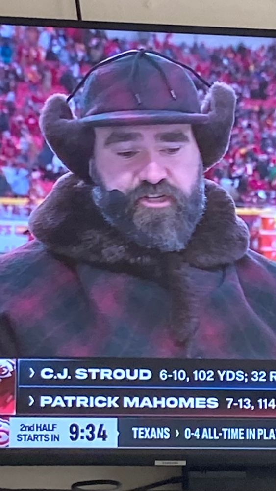 Photo of Jason Kelce on a TV broadcast wearing a red and black plaid coat with a brown fur collar and matching cap with brown fur ear flaps