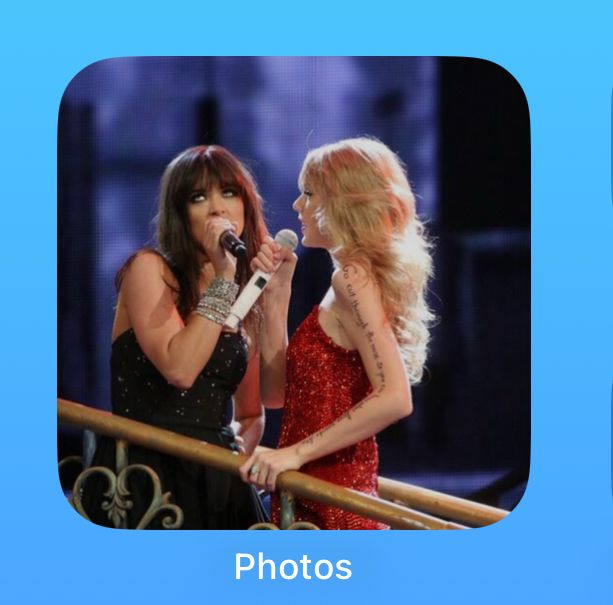 Screenshot of an iPhone Home Screen showing the photos widget and a picture of Taylor Swift in a red sequin dress with big blonde curly hair and lyrics written down her arm, gazing gayly and lovingly at Liz Huett in a black sparkly dress. They are both holding microphones