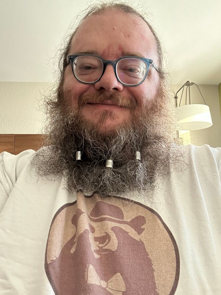 Fat, bearded white guy wearing glasses. He has beads in his beard. He’s wearing a white t-shirt with a logo of a bear in a bowler hat on it.