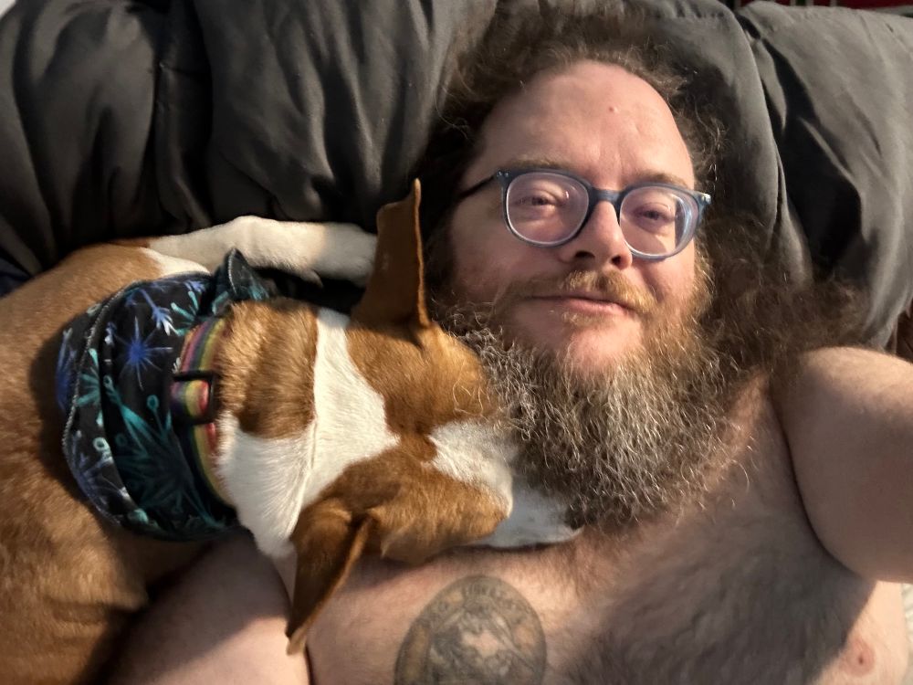 Landscape oriented photo of a shirtless man with a long white & brown beard and wearing glasses who’s laying back on a comforter. A brown and white dog lays beside him, its head resting on the man’s shoulder and neck. The shot is taken from above.