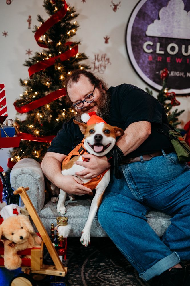 A fat white man with a beard and glasses is holding/hugging a brown and white medium-sized dog while they’re both sitting on a divan. The man is looking down at the dog and smiling. The dog is leaning into the man and smiling while looking at the camera. The dog is wearing a Santa hat. There’s a lit Xmas tree in the background.