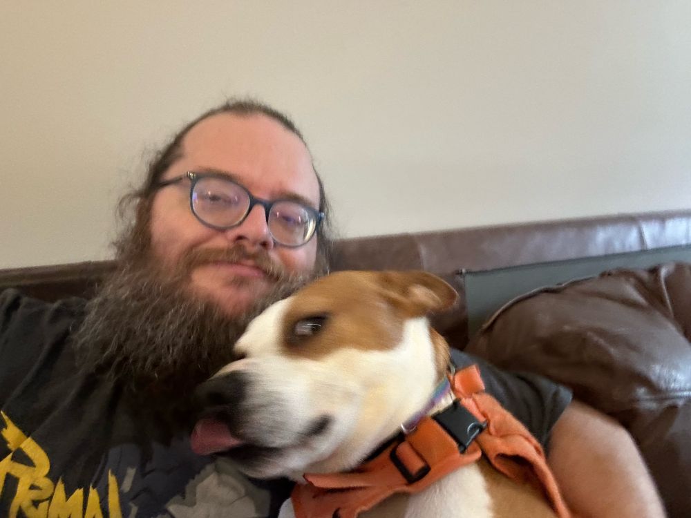Bearded white man wearing glasses sitting on a leather couch with a brown and white dog leaned against his shoulder. The dog is in 3/4 profile looking to the left and has slight motion blur as he licks his chops.