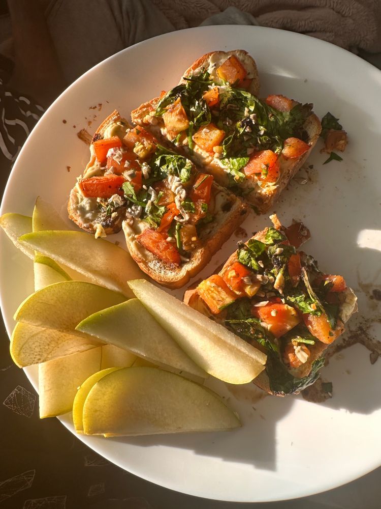 Bruschetta-inspired toast topped with tomatoes, herbs, sunflower seeds, and a deep brown glaze. Pear slices on the side. 