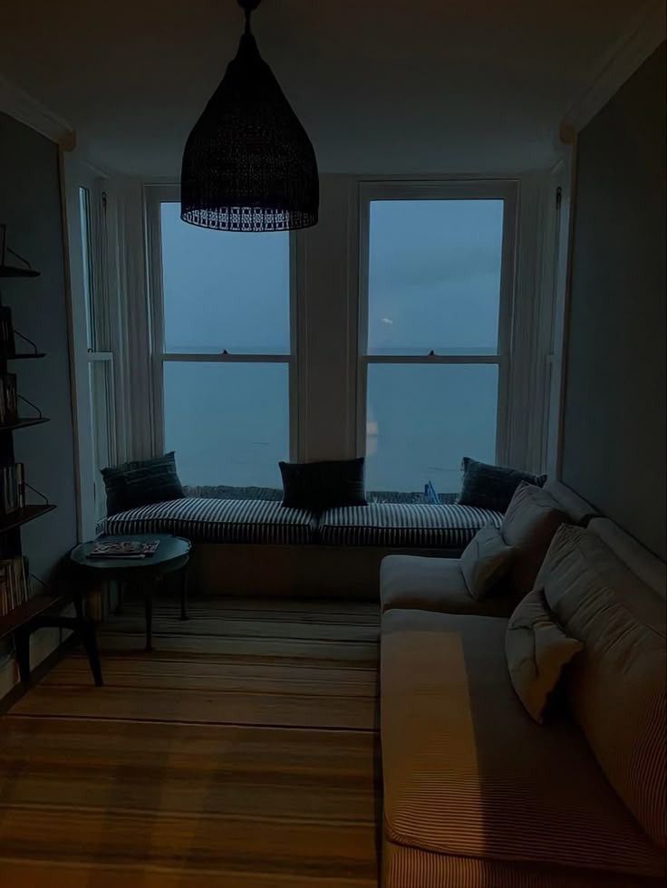 Small living room lit by a dim, warm interior light from the adjacent room and cool overcast light from the large windows on the opposite side. There is a window seat, a small glass top table, a couch, and a striped rug. A bookshelf is just barely in frame. A simple chandelier is hanging from the ceiling, currently turned off. 