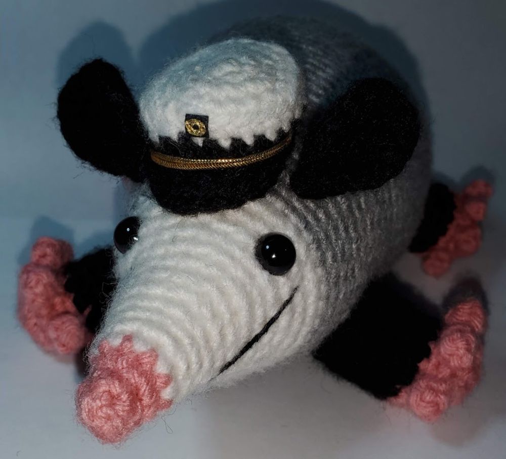 A crochet possum amigurumi done in a cutesy style wearing a crochet black and white captain's hat with gold embellishments.