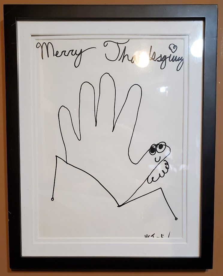 A drawing in black sharpie on white paper. At the top in cursive it reads "Merry Thanksgiving" (with a heart over the first letter i) and below that is a turkey drawn by tracing a hand and adding eyes, a smiley mouth, and a wattle to the thumb. It has spindly, weird little freak legs. It's signed by HCJustin in the bottom right (barely visible because the mat was slightly too small). An grown-ass adult made this and it's one of my most prized possessions.